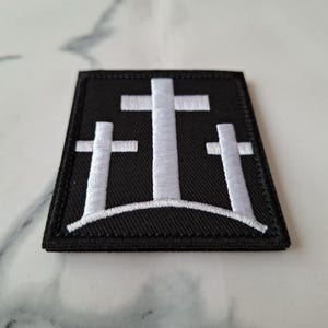 May include: A black embroidered patch featuring three white crosses on a hill. The patch has a rectangular shape with a black border and is likely designed to be sewn or ironed onto fabric.