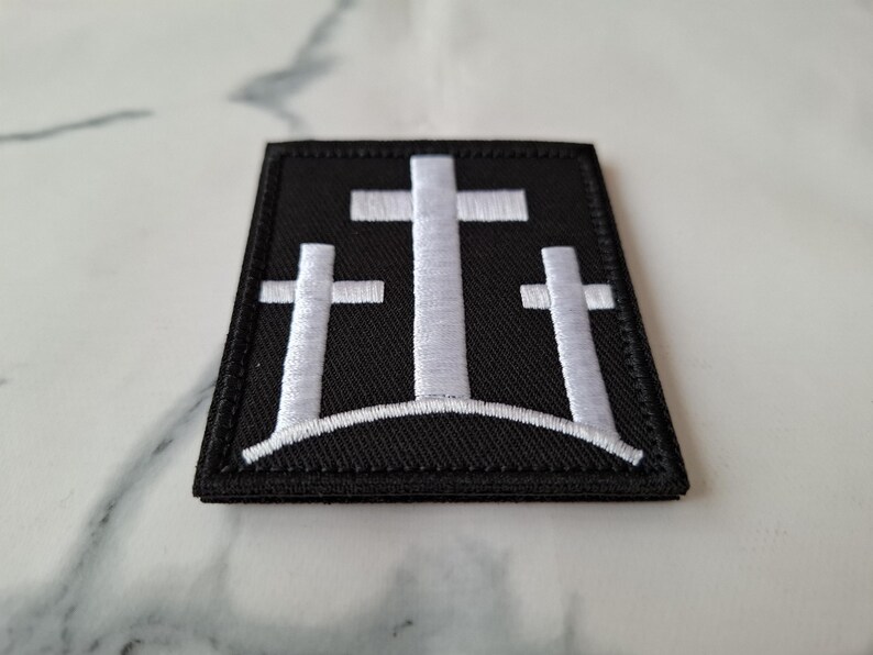 May include: A black embroidered patch featuring three white crosses on a hill. The patch has a rectangular shape with a black border and is likely designed to be sewn or ironed onto fabric.