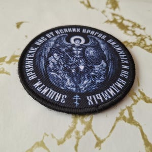 Op de afbeelding: Een geborduurde patch in zwart en wit met een afbeelding van een engel met uitgespreide vleugels. De tekst rond de engel luidt "Архистратиг Михаил - хранитель наш от всяких врагов, видимых и невидимых".