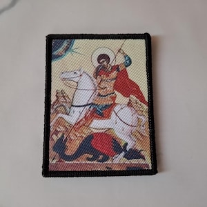 May include: Embroidered patch featuring a religious icon of a figure on horseback, spearing a dragon. The patch has a black border and depicts the rider in red and blue attire, with a white horse. The background is a light beige.