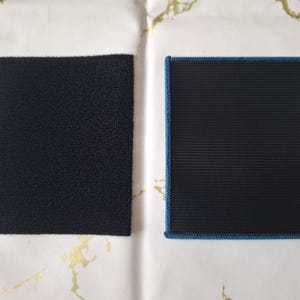 K&ouml;nnte beinhalten: Zwei schwarze Klettstoffquadrate. Eines ist ein raues, strukturiertes Rechteck. Das andere ist ein rechteckiger Aufn&auml;her mit gerippter Textur und blauem Rand. Beide liegen auf einer wei&szlig;-goldenen Oberfl&auml;che.