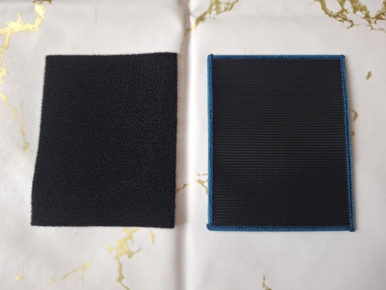 K&ouml;nnte beinhalten: Zwei schwarze Klettstoffquadrate. Eines ist ein raues, strukturiertes Rechteck. Das andere ist ein rechteckiger Aufn&auml;her mit gerippter Textur und blauem Rand. Beide liegen auf einer wei&szlig;-goldenen Oberfl&auml;che.