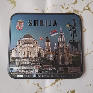 Puede incluir: Un imán rectangular con un paisaje urbano serbio, con la palabra "SRBIJA" en la parte superior. El diseño incluye una iglesia, una estatua y la bandera serbia. El imán tiene un borde negro.
