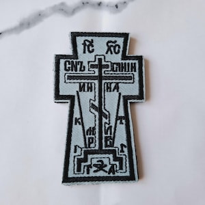 Może przedstawiać: Haftowana naszywka w kształcie krzyża z tekstem religijnym. Naszywka jest jasnoniebieska z czarnymi haftowanymi detalami i czarną obwódką. Projekt krzyża zawiera wiele linii tekstu i centralny krzyż.