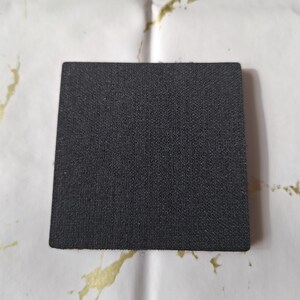 May include: A square, black coaster with a textured surface. The coaster is made of a dark material and is likely designed to absorb moisture. The background is white with gold-colored paint splatters.