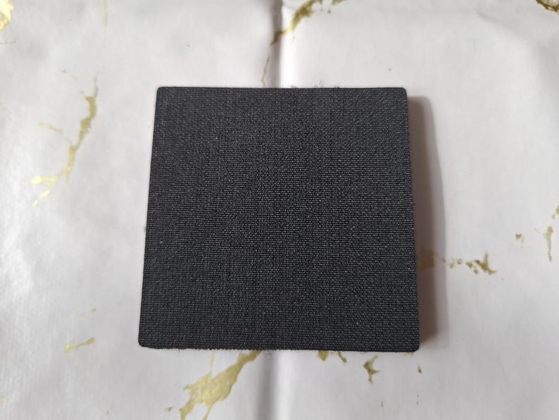 May include: A square, black coaster with a textured surface. The coaster is made of a dark material and is likely designed to absorb moisture. The background is white with gold-colored paint splatters.