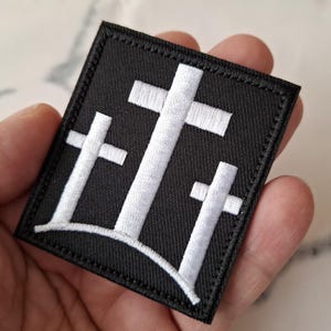 May include: Black square patch featuring three white crosses, a symbol of faith. The crosses are embroidered with a detailed, textured design. The patch has a stitched border.