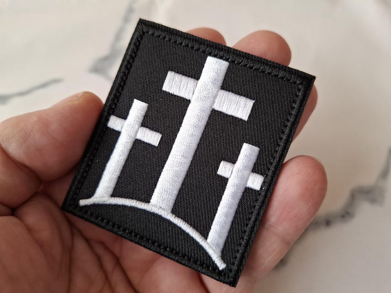 May include: Black square patch featuring three white crosses, a symbol of faith. The crosses are embroidered with a detailed, textured design. The patch has a stitched border.