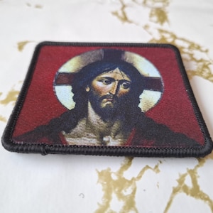 May include: A black and red embroidered patch with a depiction of Jesus Christ. The image shows Jesus with a crown of thorns and a halo. The background is red with a white halo and a cross.