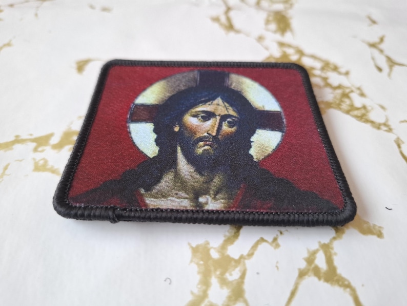 May include: A black and red embroidered patch with a depiction of Jesus Christ. The image shows Jesus with a crown of thorns and a halo. The background is red with a white halo and a cross.