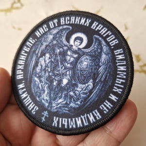 Op de afbeelding: Een geborduurde patch in zwart en wit met een afbeelding van een engel met uitgespreide vleugels. De tekst rond de afbeelding luidt "СВЯТЫЙ М. АРХАНГЕЛ,  ЗАЩИТИ НАС ОТ ВСЯКИХ ВРАГОВ, В ИМЕНИ ИХ ХРИСТА!"