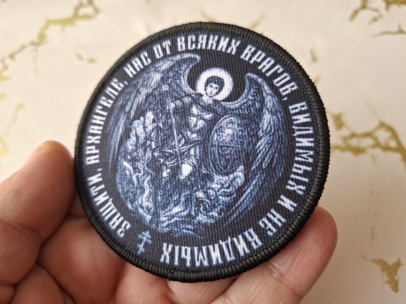Op de afbeelding: Een geborduurde patch in zwart en wit met een afbeelding van een engel met uitgespreide vleugels. De tekst rond de afbeelding luidt "СВЯТЫЙ М. АРХАНГЕЛ,  ЗАЩИТИ НАС ОТ ВСЯКИХ ВРАГОВ, В ИМЕНИ ИХ ХРИСТА!"