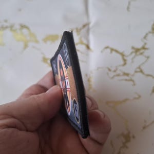 Peut inclure: Un &eacute;cusson rectangulaire avec une bordure noire. L'&eacute;cusson pr&eacute;sente un motif avec des &eacute;l&eacute;ments orange, blancs et bleus. Le motif comprend une forme circulaire avec un motif en forme de r&eacute;ticule et le chiffre 10. L'&eacute;cusson est tenu dans une main.