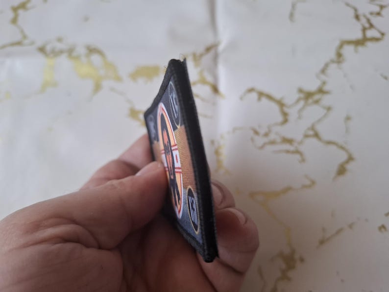 Peut inclure: Un &eacute;cusson rectangulaire avec une bordure noire. L'&eacute;cusson pr&eacute;sente un motif avec des &eacute;l&eacute;ments orange, blancs et bleus. Le motif comprend une forme circulaire avec un motif en forme de r&eacute;ticule et le chiffre 10. L'&eacute;cusson est tenu dans une main.