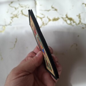 May include: A rectangular patch with a colorful illustration, featuring a black border. The patch is held at an angle, revealing its thickness. The illustration includes various colors and details, suggesting a decorative or artistic item.