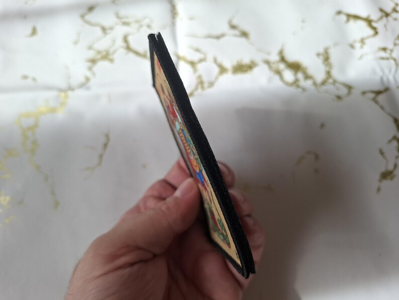 May include: A rectangular patch with a colorful illustration, featuring a black border. The patch is held at an angle, revealing its thickness. The illustration includes various colors and details, suggesting a decorative or artistic item.