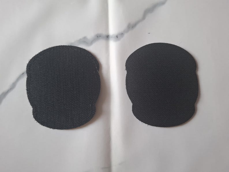 May include: Two black, skull-shaped patches with a textured surface are displayed against a white background. The patches are identical in shape and size, with a slightly rounded top and a scalloped bottom.