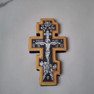 Könnte beinhalten: Ein dekoratives orthodoxes Kreuz mit einem goldfarbenen Rahmen und schwarz-weißen religiösen Bildern. Das Kreuz zeigt eine Darstellung von Jesus Christus und religiösen Text. Das Kreuz ist ein Symbol des Glaubens und der Spiritualität.