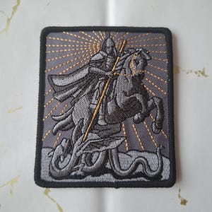 Könnte beinhalten: Gestickter Aufnäher mit einem Ritter auf einem Pferd, der eine Schlange ersticht. Das Design ist in Grautönen mit goldenen Akzenten gehalten, vor einem grauen Hintergrund mit einem Sonnenstrahlenmuster. Der Aufnäher hat einen schwarzen Rand.