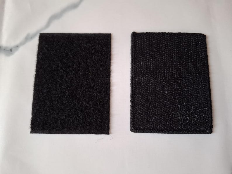 May include: Two black hook and loop fastener squares. One has a fuzzy texture, and the other has a woven texture. Both are rectangular and lie on a white surface. These are commonly used for securing items.
