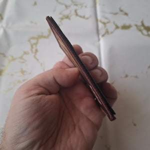 K&ouml;nnte beinhalten: Eine schlanke, braune Ledergeldb&ouml;rse wird in einer Hand gehalten. Die Geldb&ouml;rse scheint neu und unbenutzt zu sein. Die Kanten sind sauber vern&auml;ht. Der Hintergrund ist eine wei&szlig;e Oberfl&auml;che mit goldfarbener Marmorierung.