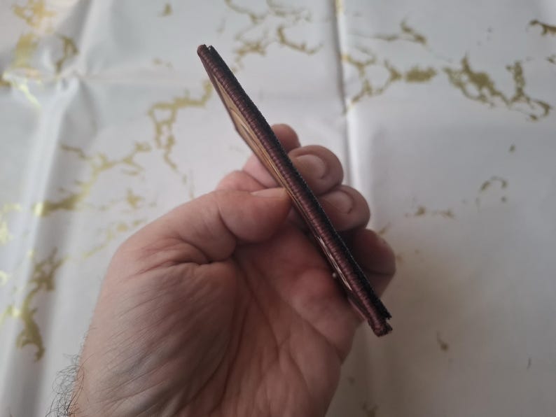 K&ouml;nnte beinhalten: Eine schlanke, braune Ledergeldb&ouml;rse wird in einer Hand gehalten. Die Geldb&ouml;rse scheint neu und unbenutzt zu sein. Die Kanten sind sauber vern&auml;ht. Der Hintergrund ist eine wei&szlig;e Oberfl&auml;che mit goldfarbener Marmorierung.