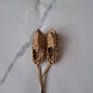May include: A pair of small, woven, brown leather shoes with laces. The shoes are tied together and are on a white background.