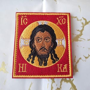 Könnte beinhalten: Gestickter Aufnäher mit einem religiösen Symbol, das eine zentrale Figur in einem goldenen Heiligenschein, dunklen Haaren und einem Bart zeigt. Das Symbol ist vor einem roten Hintergrund mit goldfarbenen Buchstaben und Verzierungen angeordnet. Der Aufnäher ist quadratisch.