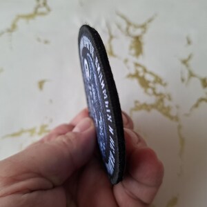 Op de afbeelding: Zwart-witte geborduurde patch met de tekst "The Reaper" en "Grim Reaper" in een cirkel. De patch heeft een zwarte rand en is gemaakt van vilt.