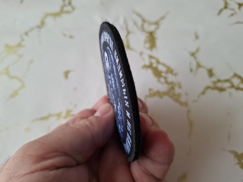 Op de afbeelding: Zwart-witte geborduurde patch met de tekst "The Reaper" en "Grim Reaper" in een cirkel. De patch heeft een zwarte rand en is gemaakt van vilt.