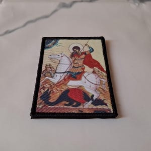 May include: Embroidered patch depicting a religious icon of a saint on horseback slaying a dragon. The patch has a black border and shows the saint in red and gold, with a white horse and a dragon in red and blue.