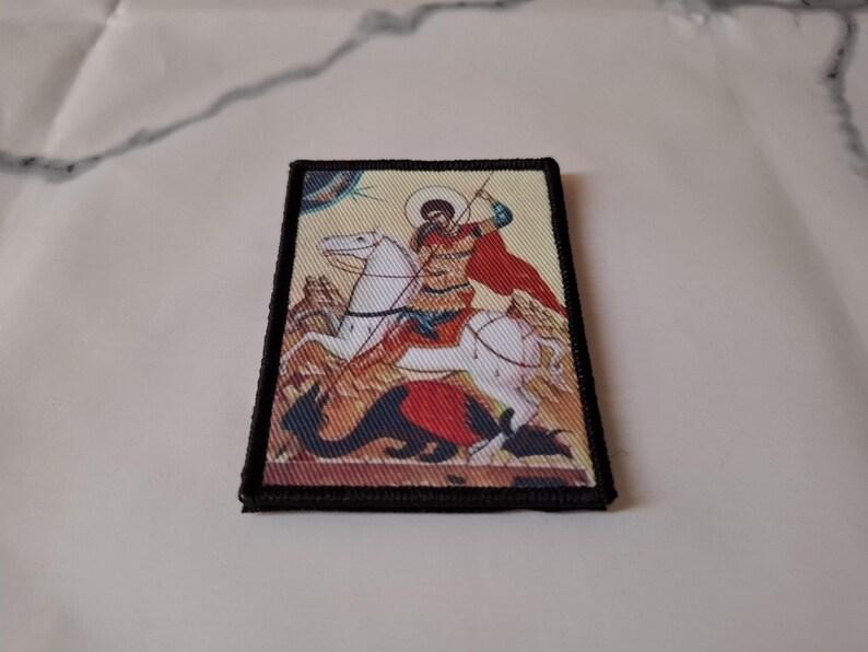 May include: Embroidered patch depicting a religious icon of a saint on horseback slaying a dragon. The patch has a black border and shows the saint in red and gold, with a white horse and a dragon in red and blue.