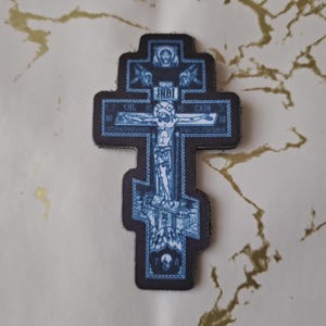 Può includere: Toppa a forma di croce ortodossa blu e nera raffigurante la crocifissione. La croce presenta dettagli e testo intricati, con un bordo. La toppa è posizionata su una superficie chiara con venature dorate.