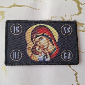Puede incluir: Parche bordado negro con una imagen colorida de la Virgen María y el niño Jesús. El parche tiene las letras "IC XC NI KA" en círculos dorados alrededor de la imagen.