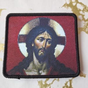 Könnte beinhalten: Ein gestickter Aufnäher in Schwarz und Rot mit einer Darstellung von Jesus Christus. Das Bild zeigt Jesus mit einem Heiligenschein und einem Kreuz hinter ihm.