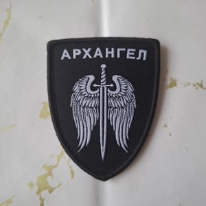 Könnte beinhalten: Schwarzer Aufnäher mit dem kyrillischen Wort "APXAHГEЛ" in Weiß. Darunter eine weiße Illustration eines Schwertes, flankiert von Flügeln. Der Aufnäher hat die Form eines Schildes.