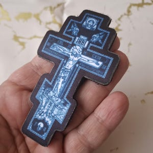 Pu&ograve; includere: Croce ortodossa blu e nera con una raffigurazione di Ges&ugrave; Cristo. La croce presenta dettagli intricati e testo, con un bordo scuro. &Egrave; un oggetto religioso.