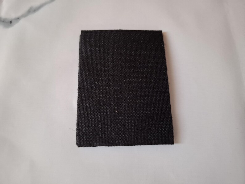 May include: A rectangular piece of black fabric with a textured weave, set against a white background. The fabric's edges are neatly cut, presenting a simple, clean appearance. The material appears to be sturdy.