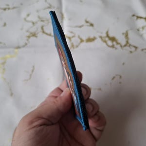K&ouml;nnte beinhalten: Ein rechteckiger Aufn&auml;her mit blauem Rand. Der Aufn&auml;her zeigt ein farbenfrohes Design mit Rot-, Orange- und Wei&szlig;t&ouml;nen. Der Aufn&auml;her wird in der Hand einer Person gehalten und zeigt sein Seitenprofil.