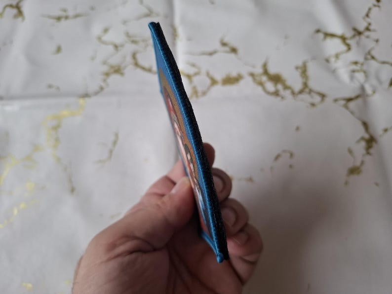 K&ouml;nnte beinhalten: Ein rechteckiger Aufn&auml;her mit blauem Rand. Der Aufn&auml;her zeigt ein farbenfrohes Design mit Rot-, Orange- und Wei&szlig;t&ouml;nen. Der Aufn&auml;her wird in der Hand einer Person gehalten und zeigt sein Seitenprofil.