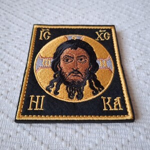 Op de afbeelding: Geborduurde patch met een religieus icoon. De centrale afbeelding is een man met donker haar en een baard, geplaatst in een gouden halo. De patch is zwart met gouden randen en letters.
