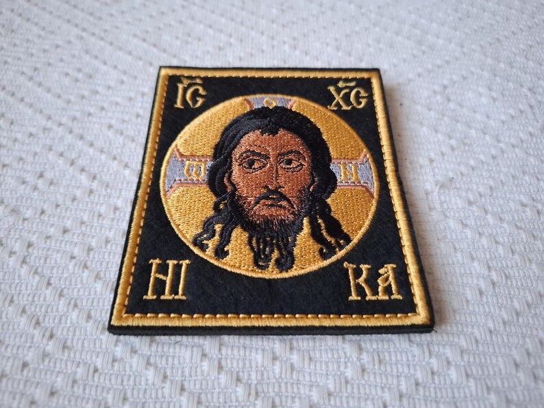 Op de afbeelding: Geborduurde patch met een religieus icoon. De centrale afbeelding is een man met donker haar en een baard, geplaatst in een gouden halo. De patch is zwart met gouden randen en letters.