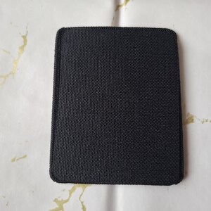 May include: A square, black fabric coaster with rounded corners. The coaster has a textured surface and is set against a white background with gold-coloured accents. The coaster is likely designed to protect surfaces from heat or moisture.