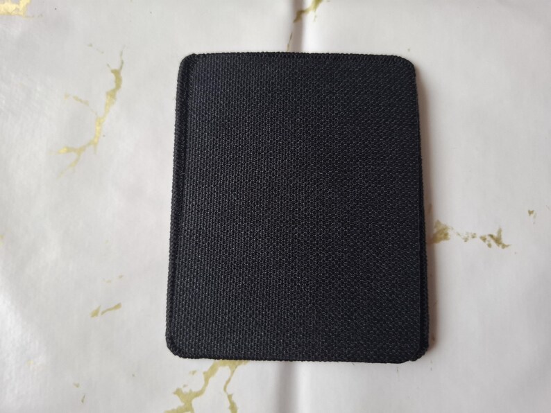 May include: A square, black fabric coaster with rounded corners. The coaster has a textured surface and is set against a white background with gold-coloured accents. The coaster is likely designed to protect surfaces from heat or moisture.