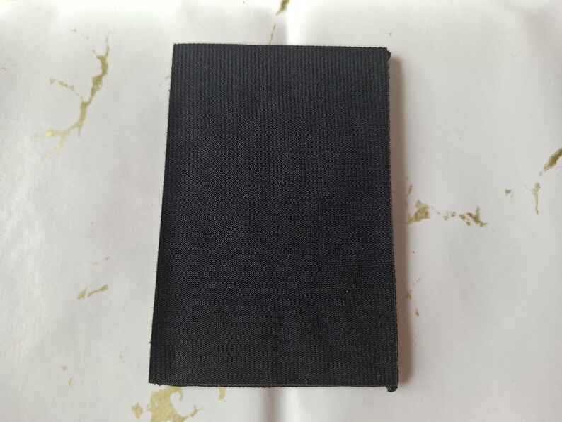 May include: A rectangular piece of black fabric with a textured, ribbed surface. The fabric is displayed against a white background with gold-colored accents. The fabric appears to be a sample or swatch.