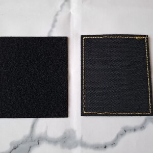 Op de afbeelding: Twee zwarte vierkante patches. De ene heeft een getextureerd, gelust oppervlak, de andere een geweven textuur met gouden stiksels langs de randen. Deze patches zijn waarschijnlijk bedoeld om aan kleding of andere items te bevestigen.