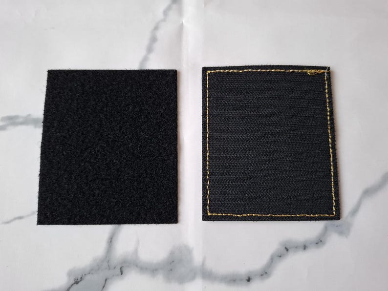 Op de afbeelding: Twee zwarte vierkante patches. De ene heeft een getextureerd, gelust oppervlak, de andere een geweven textuur met gouden stiksels langs de randen. Deze patches zijn waarschijnlijk bedoeld om aan kleding of andere items te bevestigen.