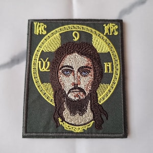 Könnte beinhalten: Gestickter Aufnäher mit einer detaillierten Darstellung von Jesus Christus. Das Bild zeigt ein Gesicht mit langen braunen Haaren, einem Bart und blauen Augen, umgeben von einem goldenen Heiligenschein mit griechischen Buchstaben. Der Aufnäher hat einen dunkelgrünen Hintergrund.