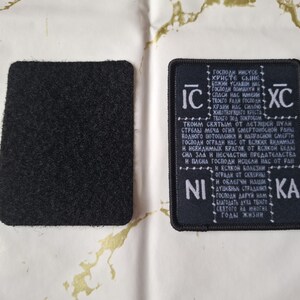 May include: Two black patches, one embroidered with white text and symbols, the other a plain square. The text is in a foreign language, possibly religious. The patches are on a white surface with gold patterns.