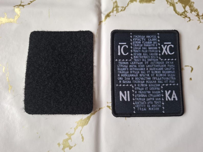 May include: Two black patches, one embroidered with white text and symbols, the other a plain square. The text is in a foreign language, possibly religious. The patches are on a white surface with gold patterns.